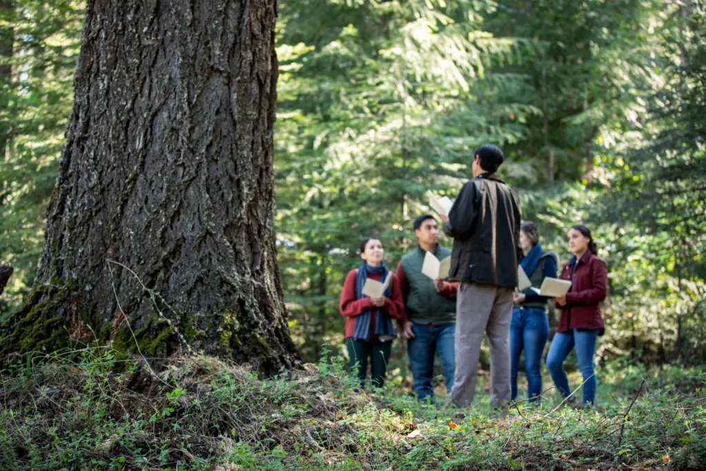 A group is gathered for a memorial of a loved one at the base of their memorial tree. They hold programs with memories and poems to honor their lost family member.