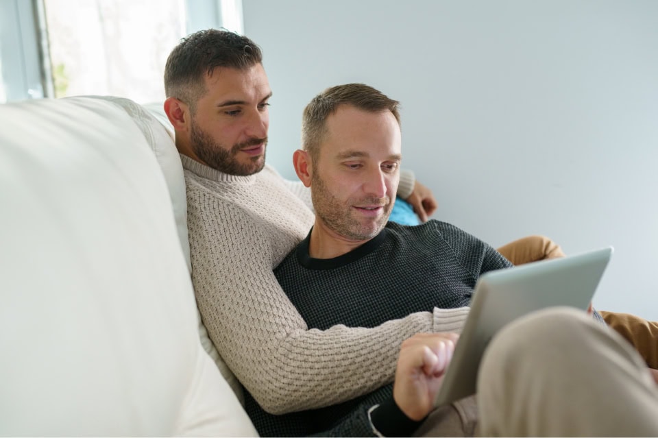 Two men sit closely together on a white couch, both looking at a laptop.