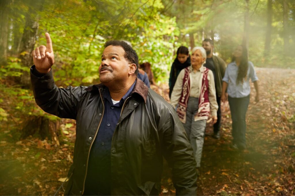A man points upward toward the tree canopy on a forest path, with a group of people walking behind him.