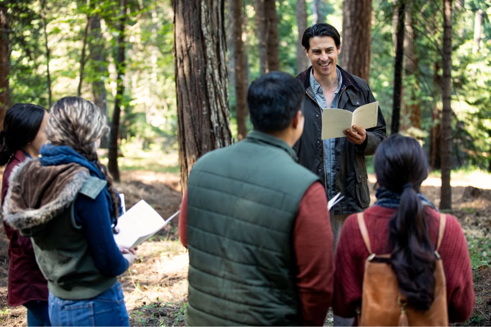 A man smiles while holding a booklet and speaking to a small group in a sunlit forest.