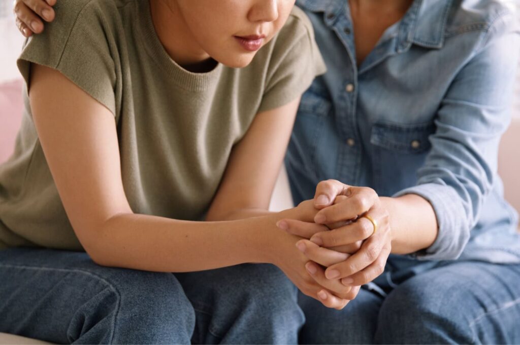 Two people sitting side by side, one gently holding the other's clasped hands in a gesture of support.